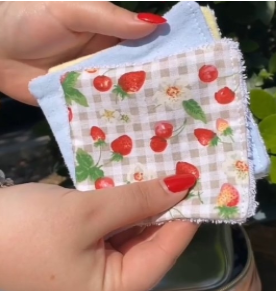 Main tenant une lingette démaquillante réutilisable à motif de fraises sur fond à carreaux, accompagnée d’une lingette bleue unie. Produit lavable, écologique et fait main, idéal pour une routine beauté zéro déchet.